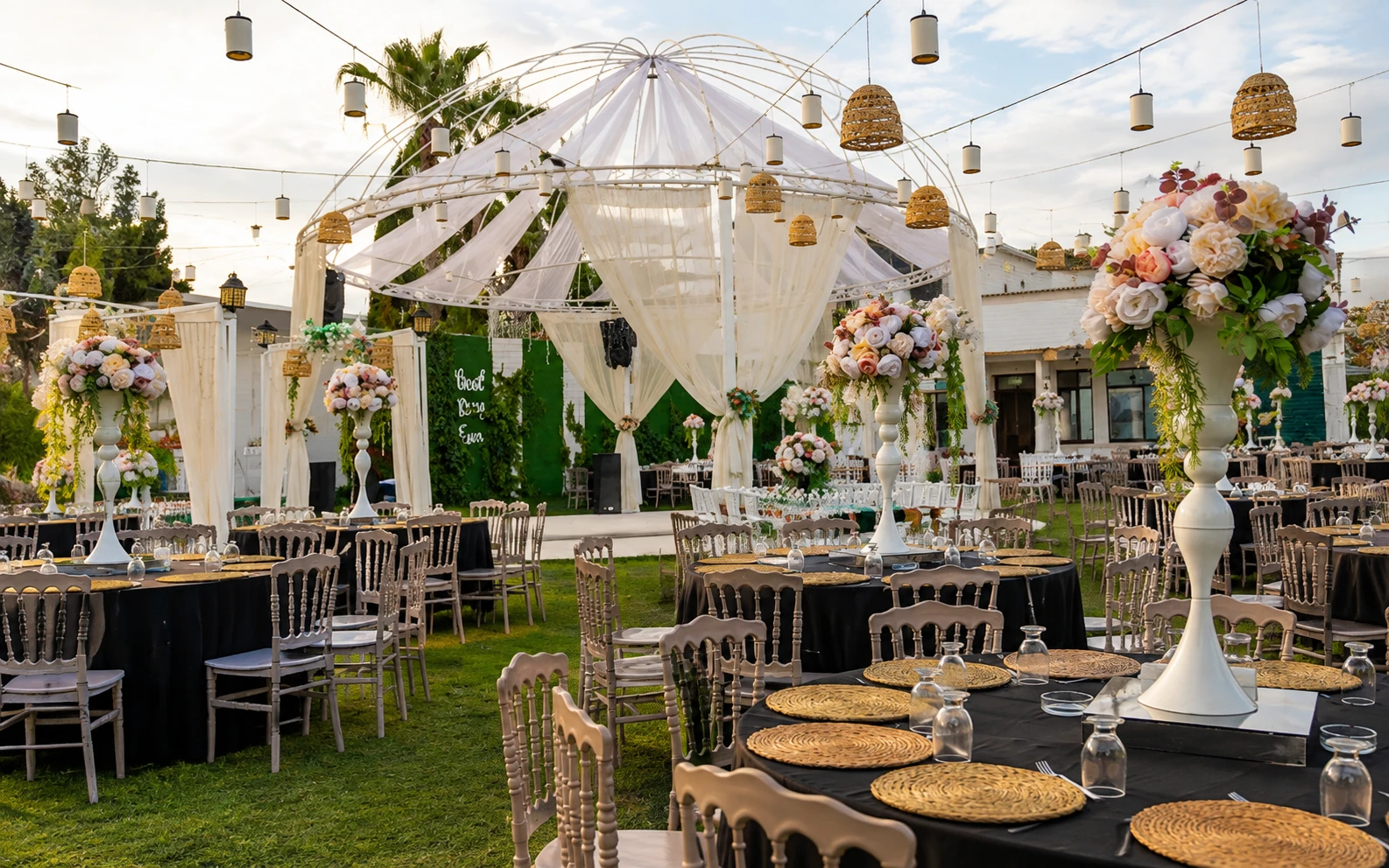 Garden wedding reception under a white draped canopy on a metal frame, with hanging lanterns, tall white floral urns with pink and cream arrangements, and round tables set on the lawn