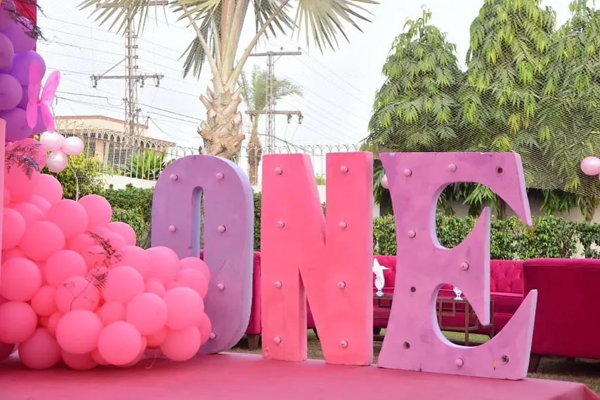 First-birthday setup with large pink and lilac ONE marquee letters beside a pink balloon garland — Baraka Events, Lahore