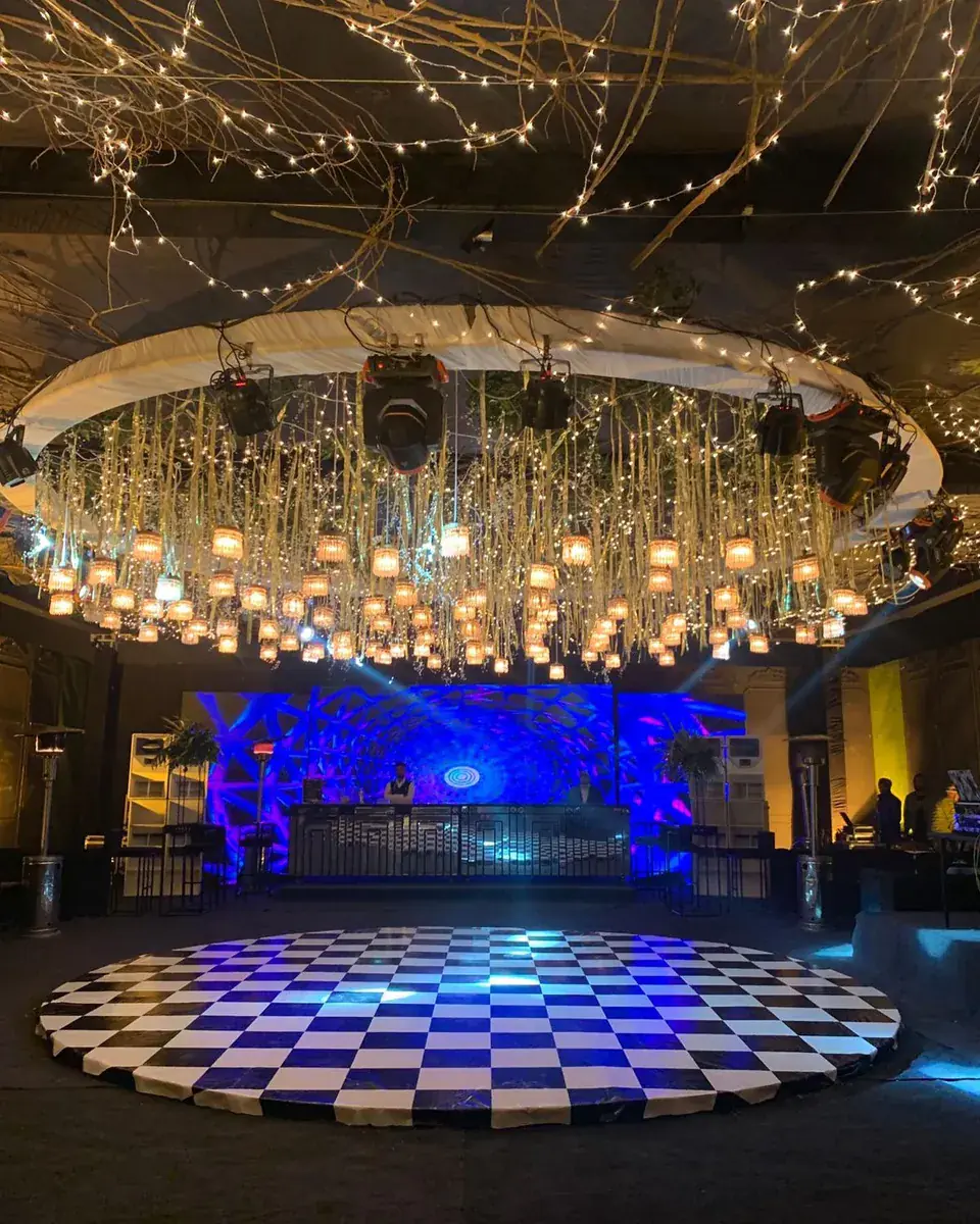 Round checkerboard dance floor beneath a ring of hanging lanterns and moving lights facing a DJ booth