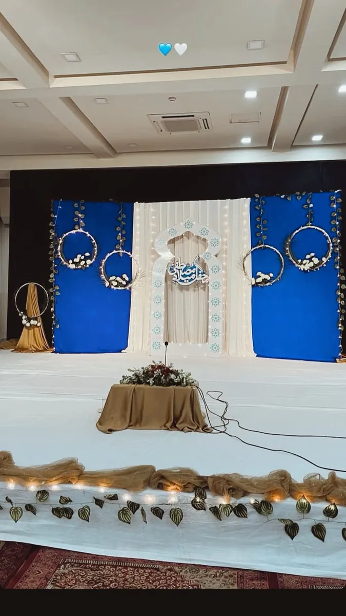 Mehfil-e-milad stage with blue panels, white-flower hoops and a lit arch with Arabic calligraphy