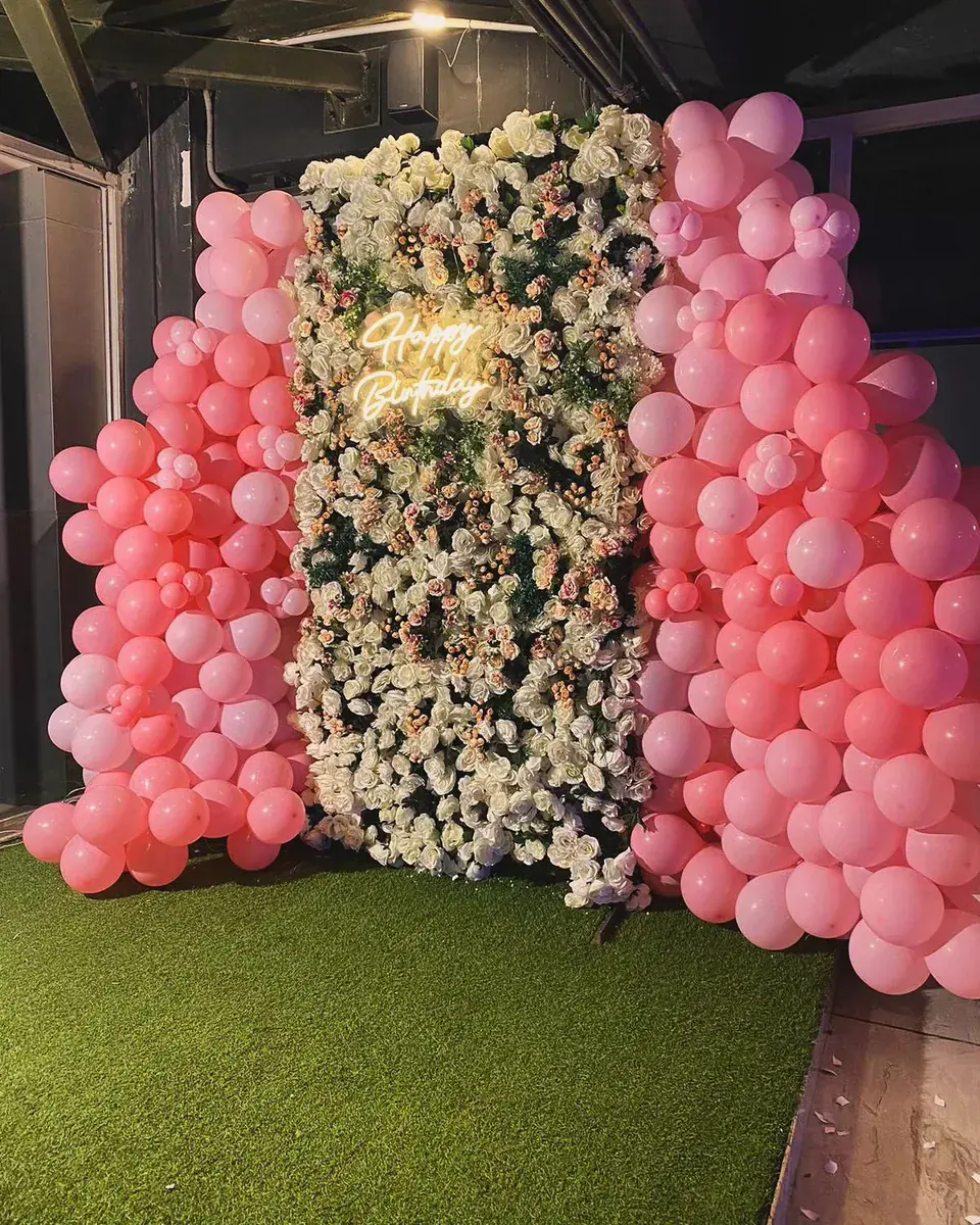 White flower wall with a Happy Birthday neon sign between two pink balloon columns