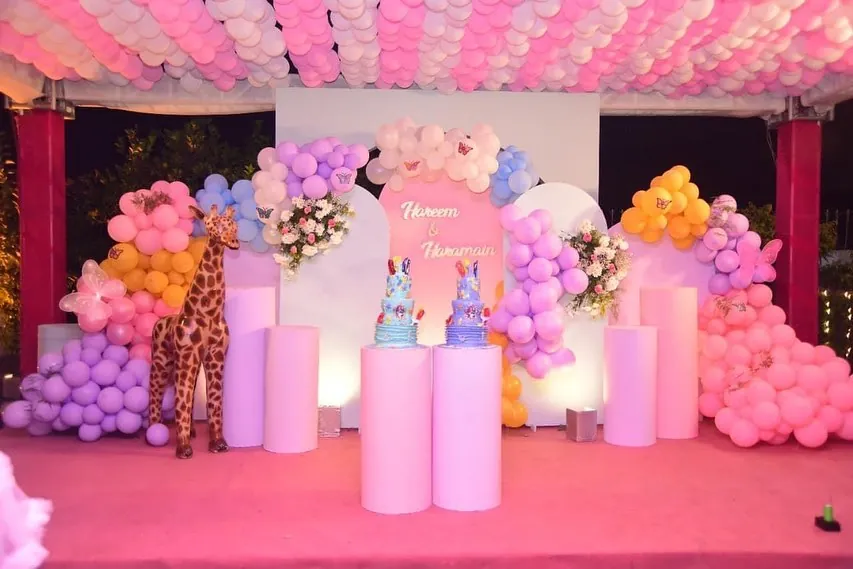 Birthday stage with a pastel balloon backdrop, giraffe figure and two tiered cakes on pink stands