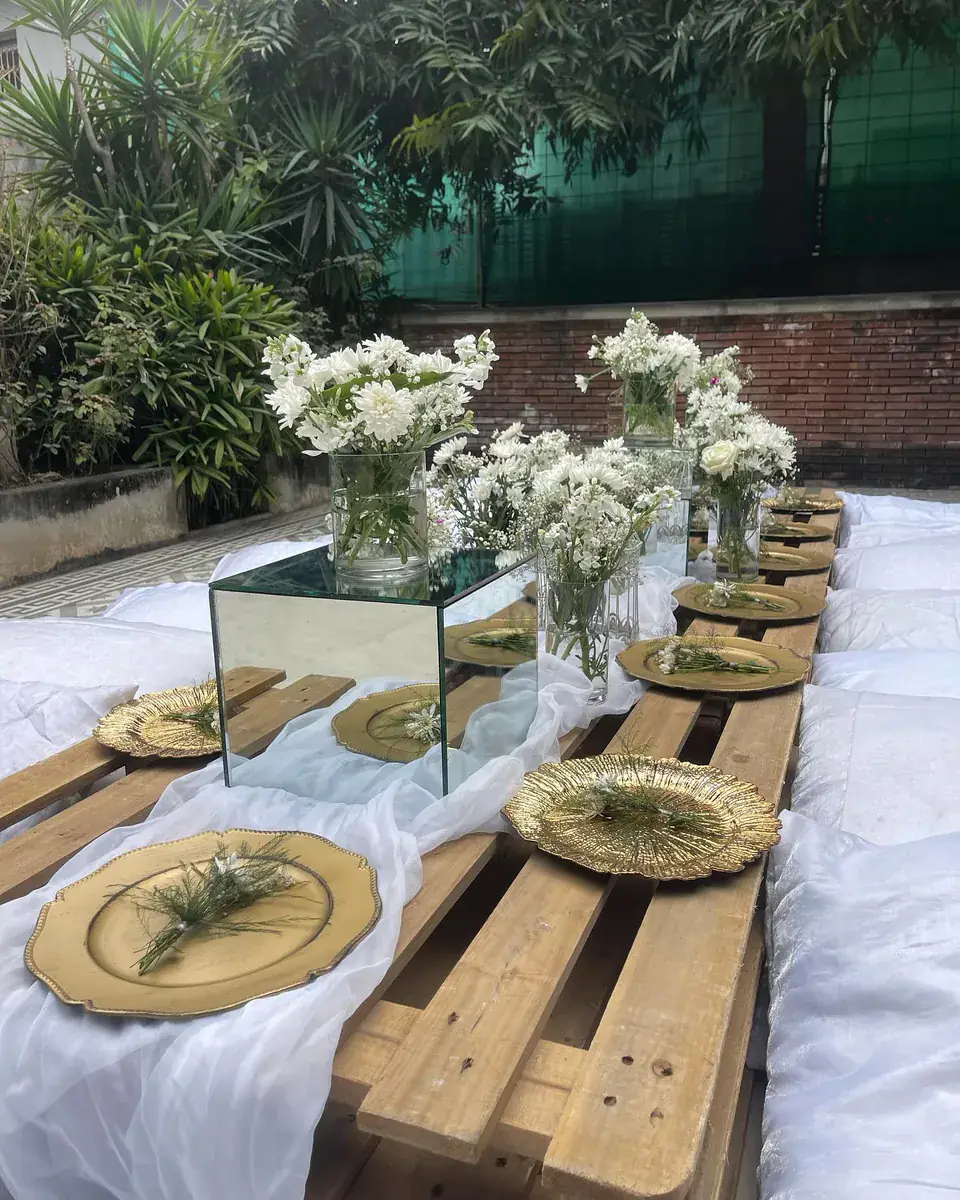 Picnic-style party table made from wooden pallets with gold plates, white flowers and a mirrored cube