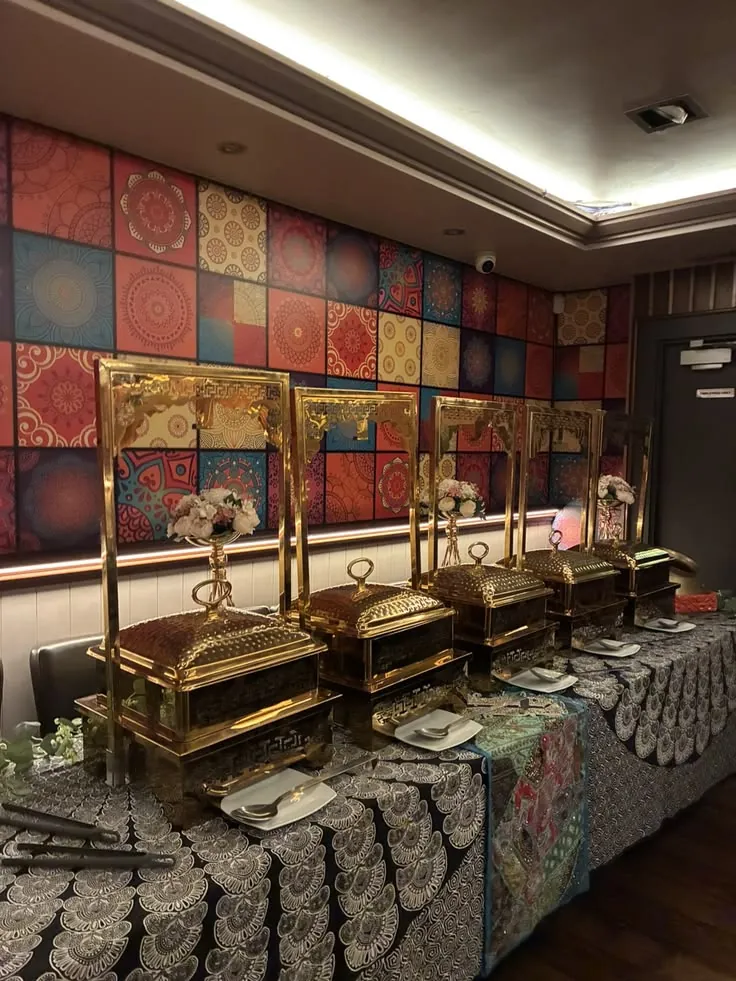 Buffet line of gold-framed serving dishes with small flower arrangements on patterned table linen