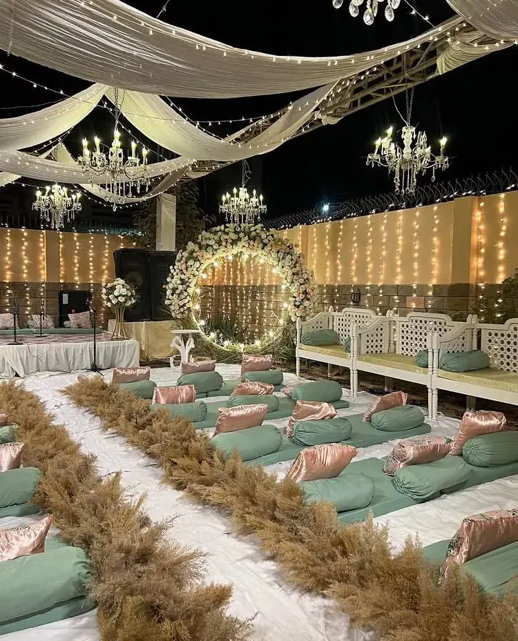 Floor seating with sage-green bolsters, pink satin cushions and a flower hoop arch under chandeliers