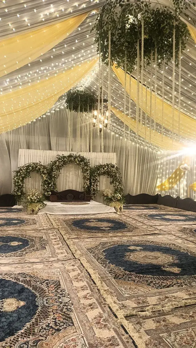 Tent interior for a qawali night with patterned carpets, a leafy floral-arch stage and a fairy-light draped ceiling