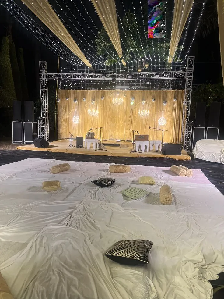 Open-air live-music stage with a gold curtain backdrop, globe chandeliers and microphones facing floor seating