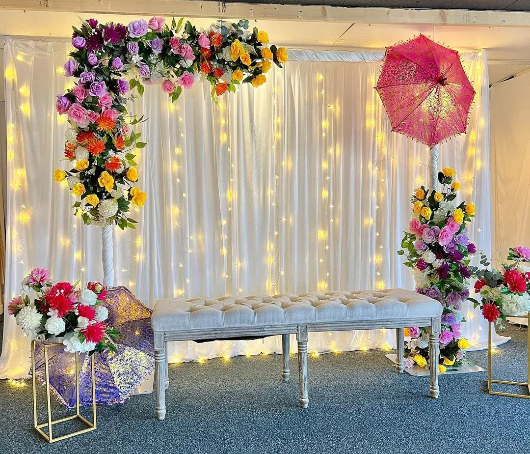 Floral arch of pink, orange and purple flowers beside a tufted bench and a pink lace parasol — Baraka Events, Lahore