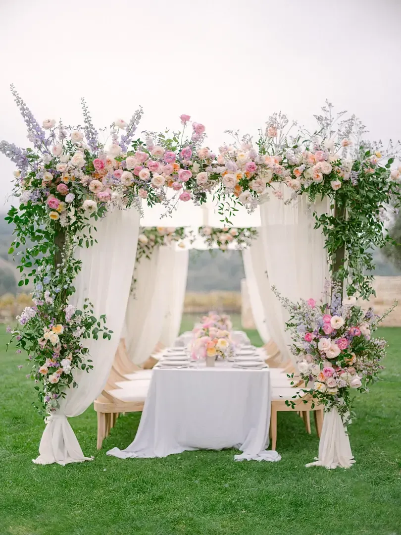 Pastel floral arches with sheer drapes over a long dining table on a lawn