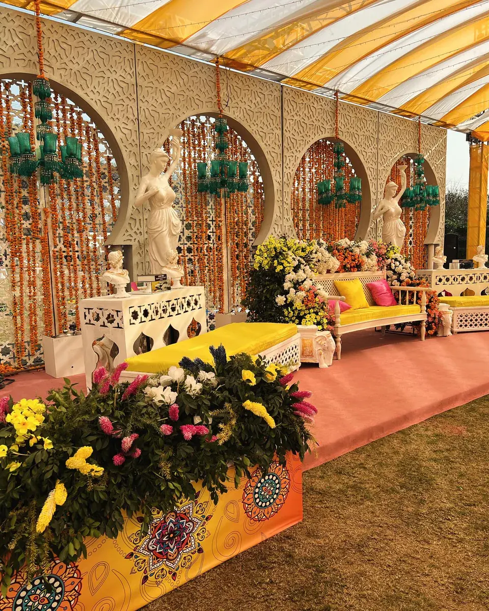 Mehndi stage of carved arches hung with marigold garlands, white statues and yellow cushions — Baraka Events, Lahore
