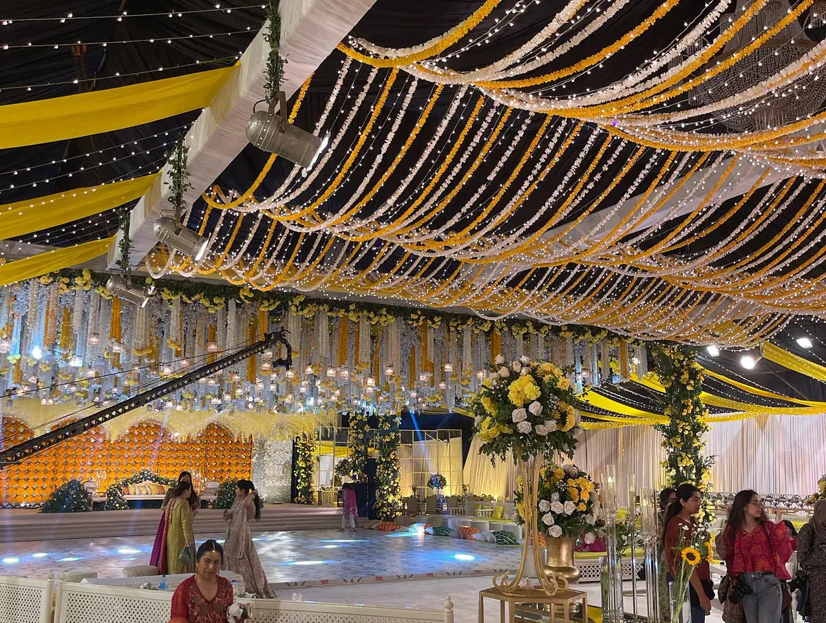Mehndi hall with a fairy-light ceiling, yellow and white garlands and a marigold stage wall beyond flower urns