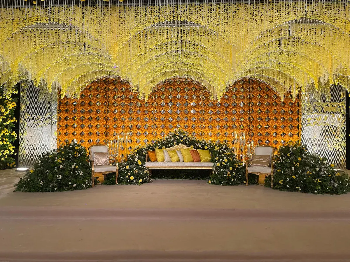 Mehndi stage with cascading yellow flower strands, a marigold backdrop and a white sofa between flower mounds