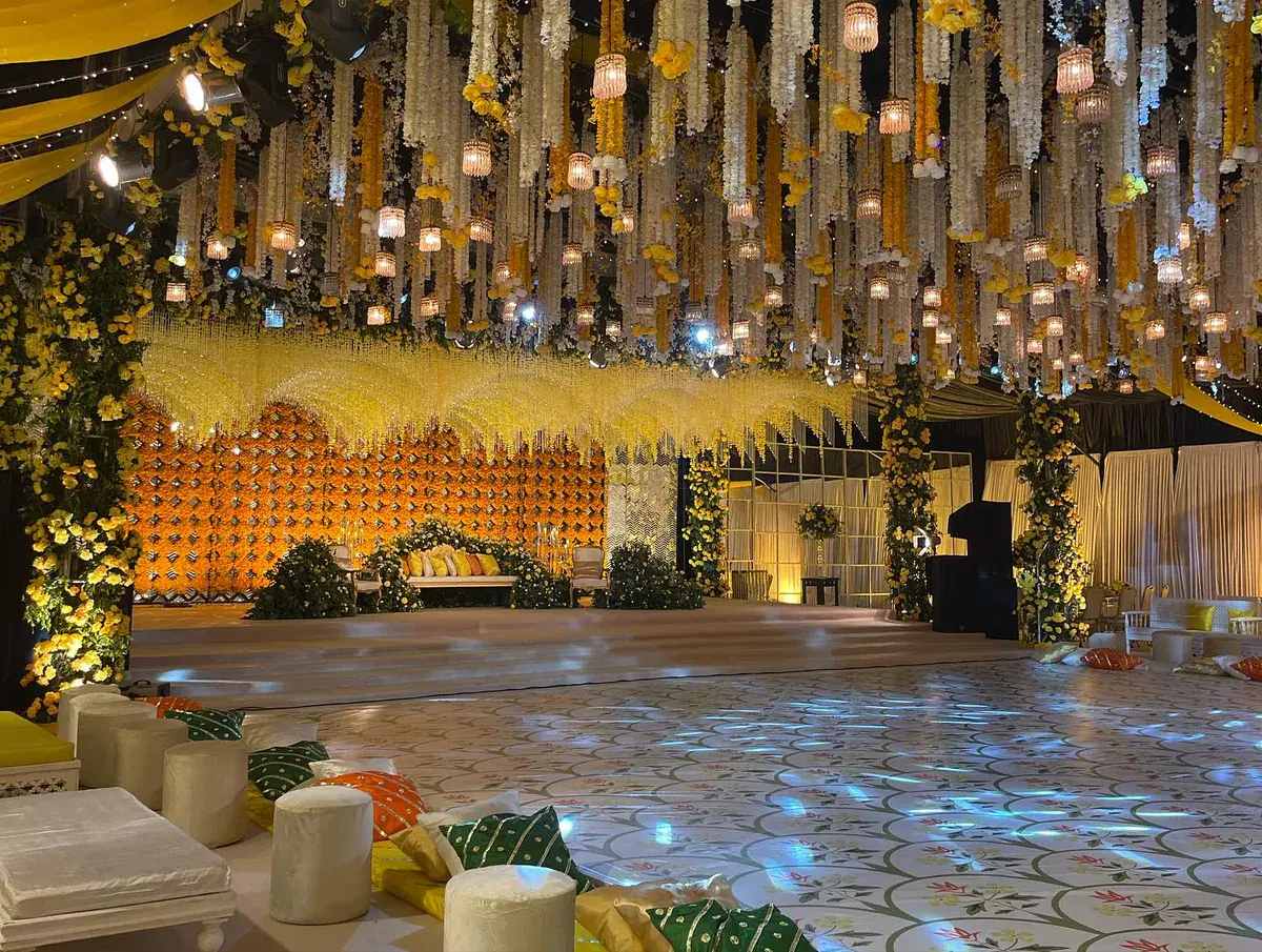 Mehndi ceiling of hanging garlands and lanterns above a marigold stage wall, with floor cushions on a lit floor