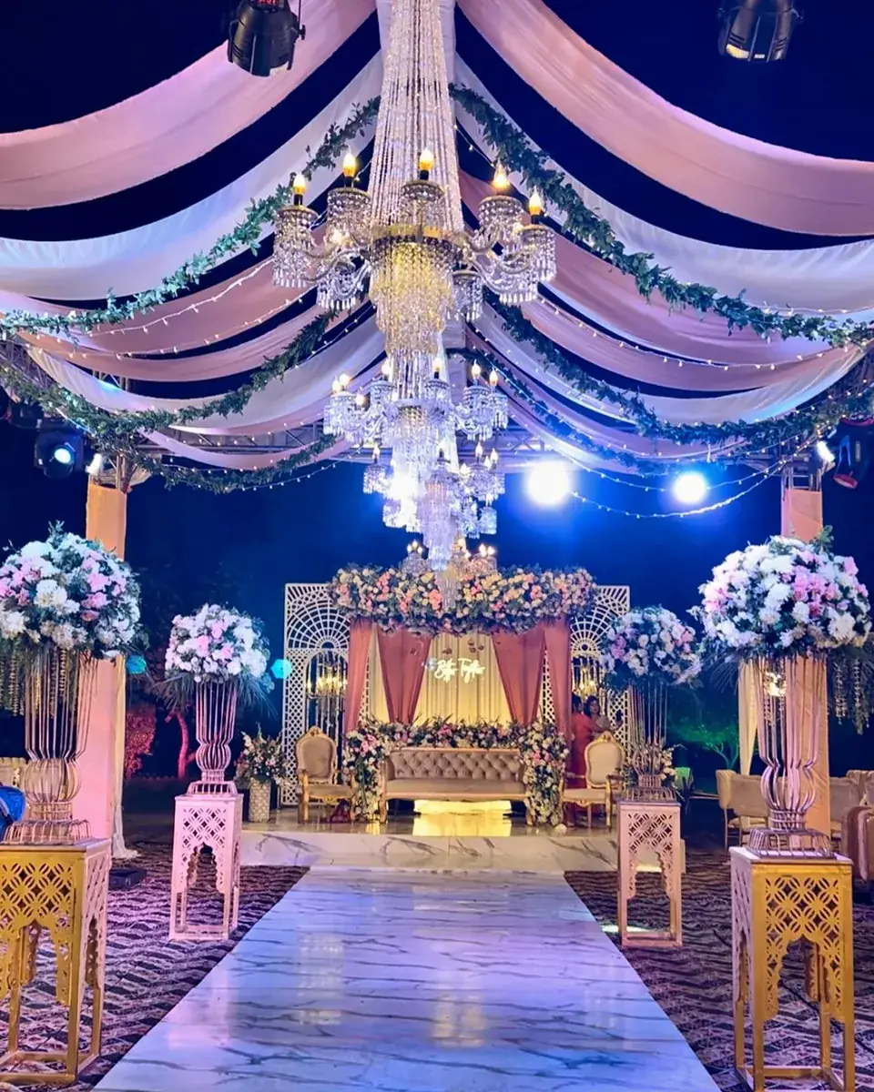 Evening wedding stage under a draped pink and white ceiling with a crystal chandelier and tall floral urns