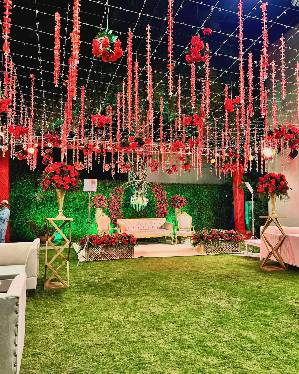 Mehndi lawn with red flower garlands hanging from a fairy-light grid and a gold sofa stage against a green hedge