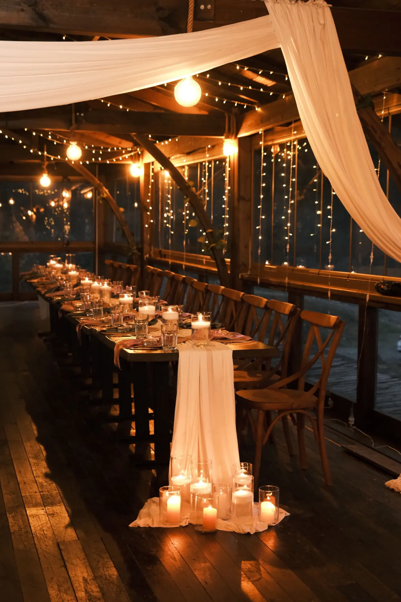 Long candlelit dining table in a timber pavilion, with white draped fabric overhead, string lights, a cream table runner and candles in glass holders on the floor