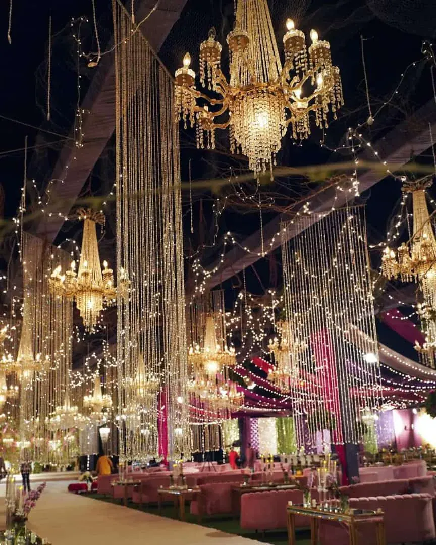 Rows of hanging crystal chandeliers and bead curtains over a pink lounge beside a white aisle at night