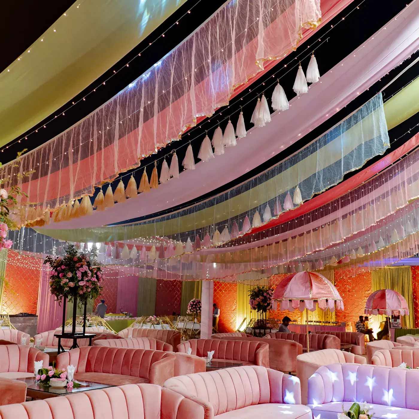 Pastel draped ceiling with tassels and fairy lights above pink velvet lounge sofas and parasols