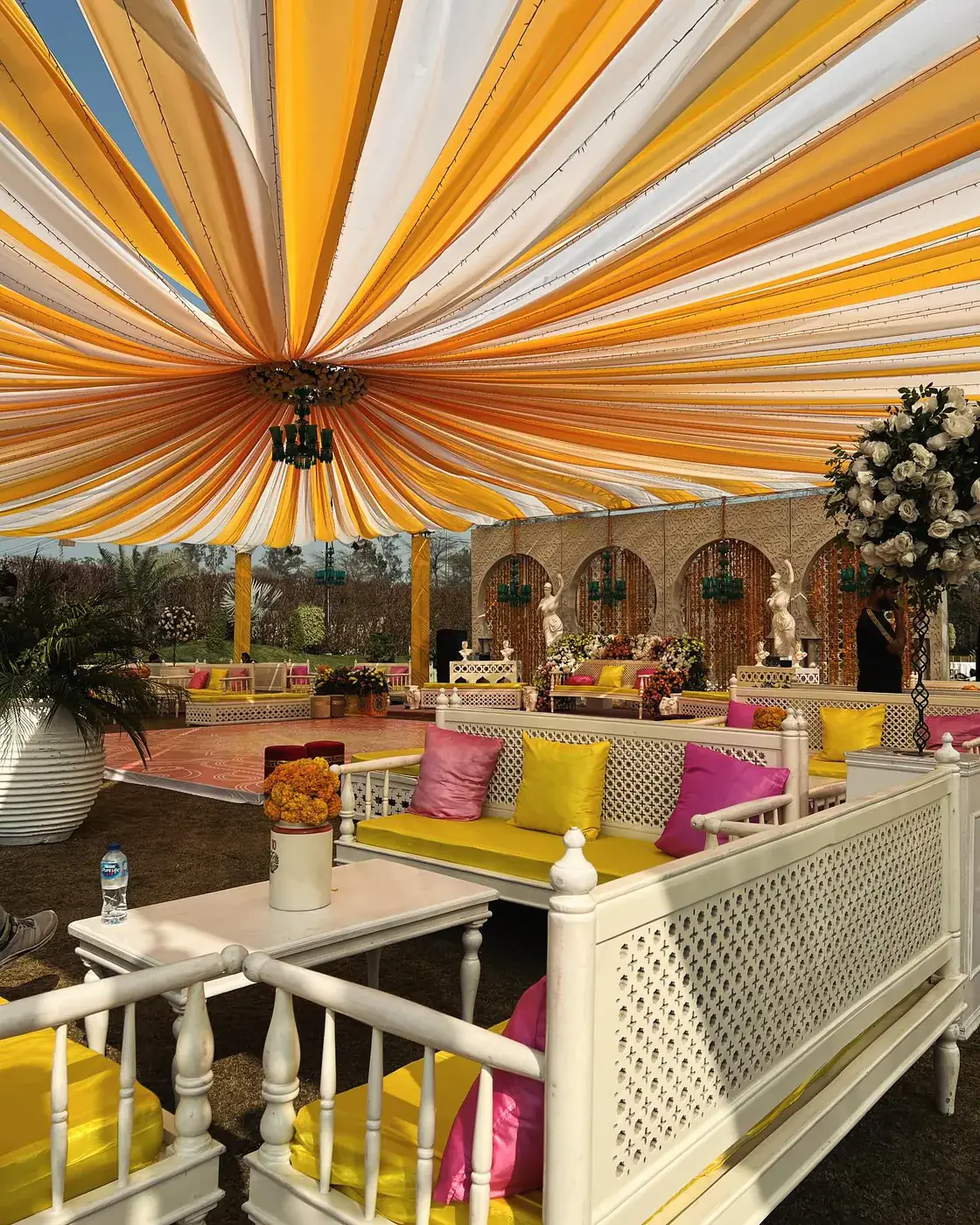 Orange and white draped canopy over white lattice sofas with yellow and pink cushions in daylight